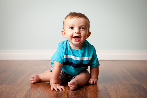 Color image of a happy six-month-old baby boy smiling while sitting on a shiny hardwood floor, with light blue background.