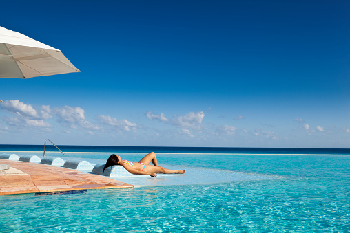 Young woman on a tourist resort vacation, relaxing by a tropical beach resort hotel infinity pool on the Caribbean Sea, Cancun, Riviera Maya, Mexico.