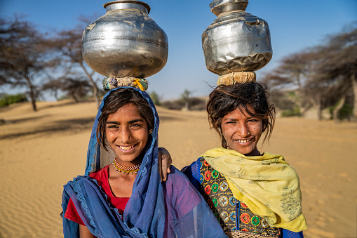 Indian young girls crossing sand dunes and carrying on their heads water from local well, Thar Desert, Rajasthan, India. Rajasthani women and children often walk long distances through the desert to bring back jugs of water that they carry on their heads.