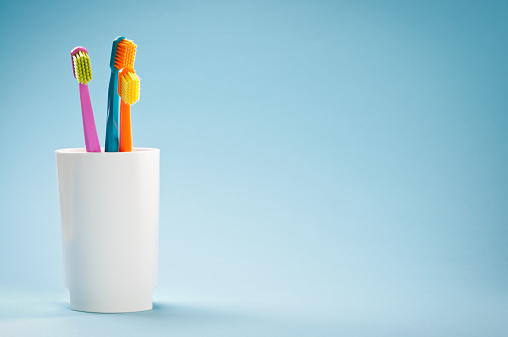 Three colourful toothbrushes in mug on light blue background. Copy space, studio shot.