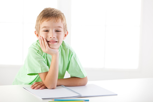 Happy little boy sitting by the table, leaning on opened notebook, day dreaming.