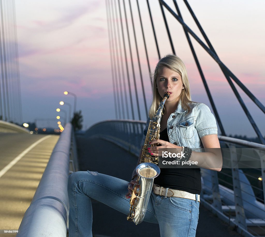 Joven mujer tocando el saxofón en iluminado puente de tirantes al atardecer - Foto de stock de 20-24 años libre de derechos
