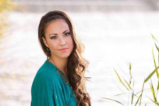 Portrait of beautiful young brunette woman, wearing elegant blue dress, standing on a stone river bank with green trees and blue sky on background