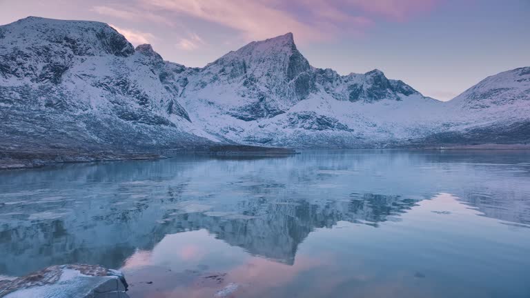 Aerial view from low flying drone of sea shore, snowy mountains in winter at sunset. Lofoten islands, Norway. Top view of fjord, reflection, water, stones, snowy rocks, purple sky with clouds. Lake