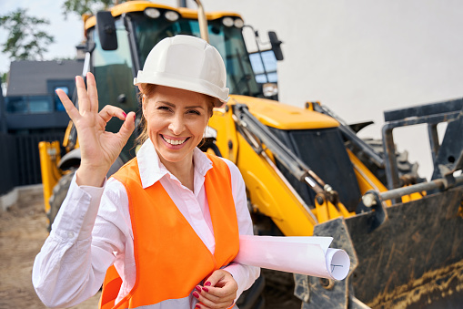 Waist-up portrait of cheerful building inspector making OK sign while standing near backhoe loader