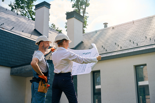 Joyous contractor pointing at roof of new house while looking at architectural drawings in construction manager hands