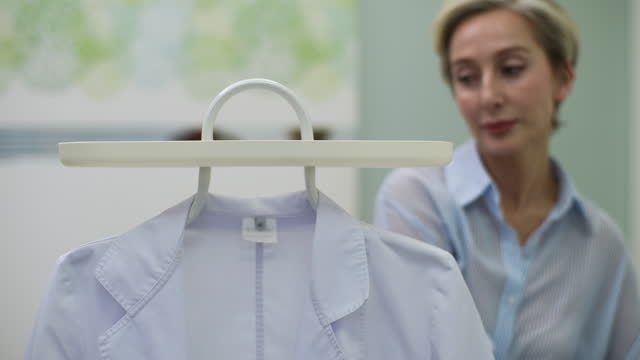 Serious middle-aged female doctor walks into office, takes robe off coat rack and puts it herself. Portrait of senior aged medical practitioner