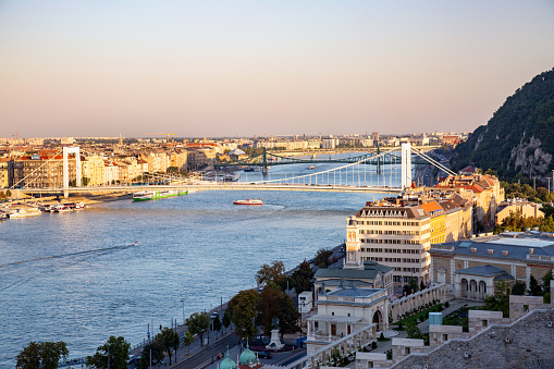 The Elizabeth Bridge in Budapest, Hungary. The bridge is a white suspension bridge with blue cables. The bridge spans the Danube River. The photo is taken from a high vantage point, looking down on the bridge and the river. The photo is taken at sunset, with the sun casting a warm glow on the buildings and the bridge. The buildings on either side of the river are a mix of modern and historic architecture. The river is busy with boat traffic.