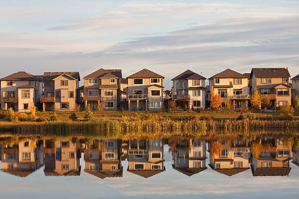 Residential Homes and Neighborhood in Suburbs Calgary Alberta A row of houses reflected in a pond. Horizontal colour image. This classic new neighbourhood or residential region in a large North America City - this is Calgary, Alberta - speaks to the urban sprawl as millions of detached homes cover massive acres or urban area. Here a beautiful wetlands region is close to these homes and provides a nice morning reflection.  alberta stock pictures, royalty-free photos & images