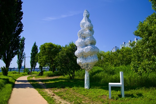 The 'Han River Tree-P6', a 7.2-meter pixelated mulberry tree sculpture, stands next to a walking path, symbolizing the limits of human perception, accompanied by an oversized white chair.