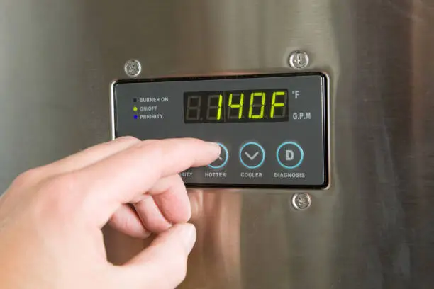 "A male finger is turning up the temperature on a tankless water heater control panel. Tankless water heaters are new technology which produces hot water on demand and is much more energy efficient than a conventional hot water holding tank system. This unit is installed and in operation, there are some scratches, dust and scuff marks on the surface."