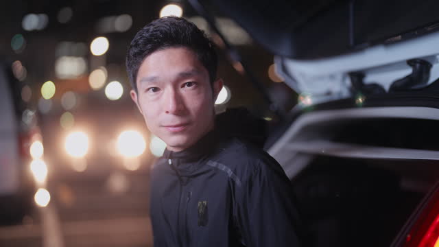 Portrait of male  Asian athlete sitting on his car trunk and getting ready for training and running in parking lot in city at night