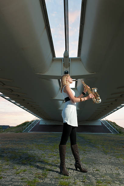 full length side view shot of a young blond woman playing on saxophone standing under bridge at sunset