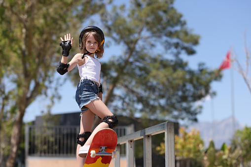 Teenage girl in helmet standing on skateboard against blue sky outdoors. Teaching children to skateboard