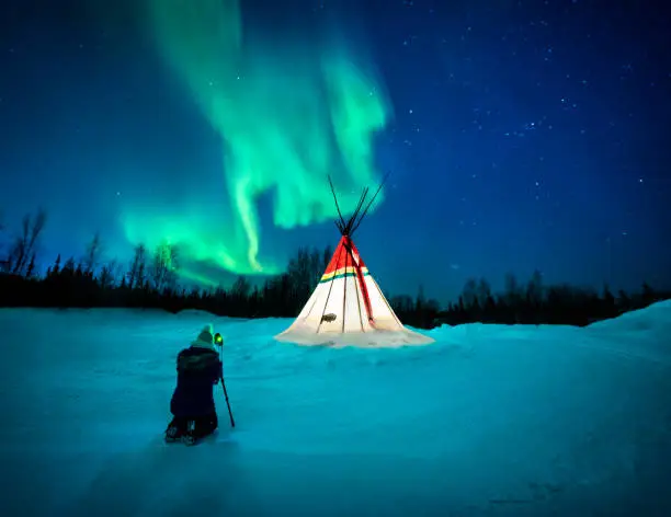 Female kneeling with camera on a tripod photographing the aurora borealis above an illuminated teepee, Yellowknife, Northwest Territories, Canada