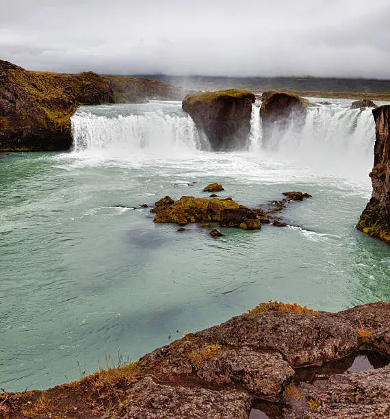 "Square image of GoAafoss (falls), near Akureyri, Iceland, with glacial water spilling into the chasm below.  A large rock outcrop can be seen in the foreground."