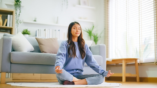 Happy young Asian woman practicing yoga and meditation at home sitting on floor in living room in lotus position and relaxing with closed eyes. Mindful meditation and wellbeing concept