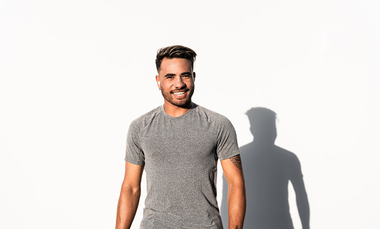 Confident young man with positive emotion standing next to white wall looking at camera.