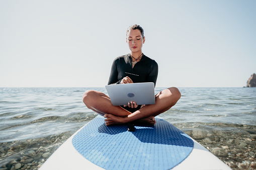 Digital nomad, freelancer, laptop. Happy smiling woman working on sup board at calm sea beach, relieves stress from work. Freelance, remote work on vacation, travel and holidays concept.