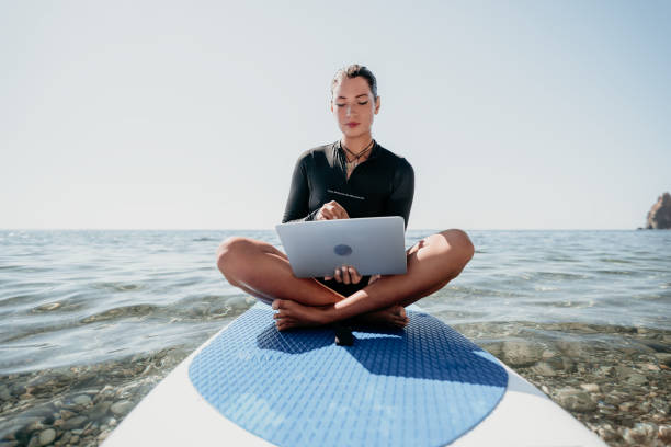 Digital nomad, freelancer, laptop. Happy smiling woman working on sup board at calm sea beach, relieves stress from work. Freelance, remote work on vacation, travel and holidays concept. Digital nomad, freelancer, laptop. Happy smiling woman working on sup board at calm sea beach, relieves stress from work. Freelance, remote work on vacation, travel and holidays concept. relieves stock pictures, royalty-free photos & images