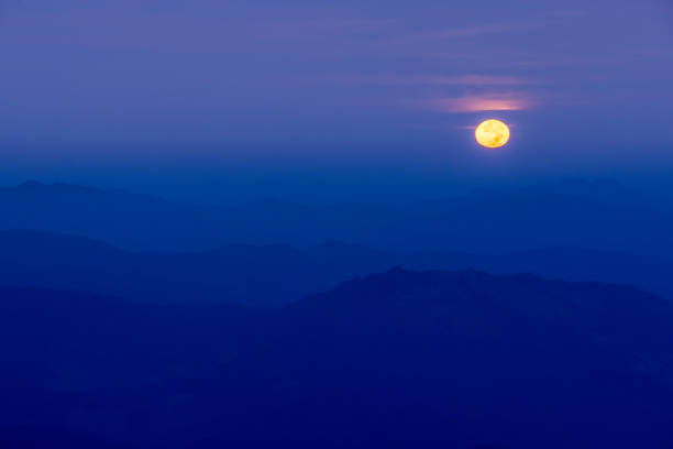 The full moon shines over blue mountains at dusk, the full moon against a dark blue sky in the background.