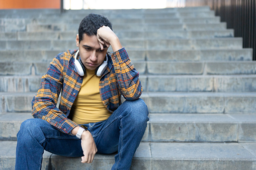 Worried and sad latin young man sitting on uran stairs with hand on the head thinking