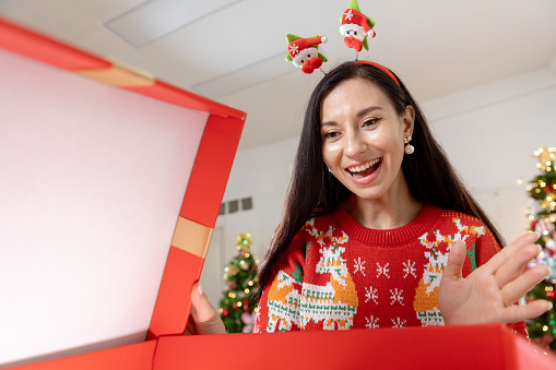 Christmas day concept. woman sitting surprised and opening gift box with christmas tree background. Festive atmosphere party.