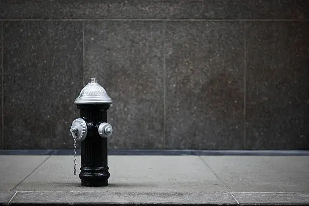 Black and Silver Fire Hydrant on New York City Sidewalk and in front of a granite wall.Sharp focus is on the Hydrant.