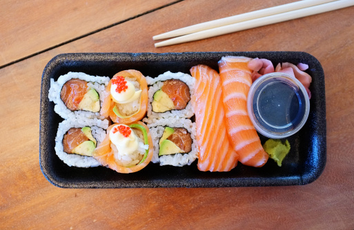 Sushi take-out containing a selection of salmon sushi specialities and sauces in a polystyrene tray with chopsticks on the side.