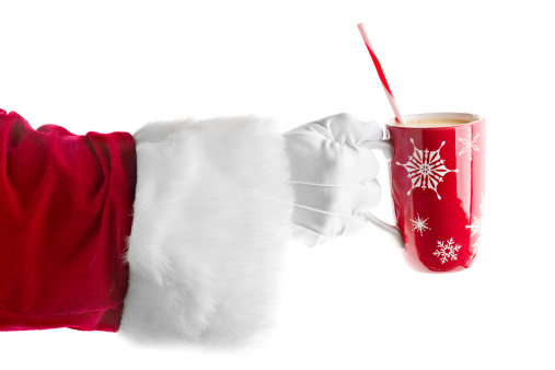 A close up of Santa's hand holding or offering a holiday decorated red mug full of delicious eggnog with a red and white straw. Isolated on white.