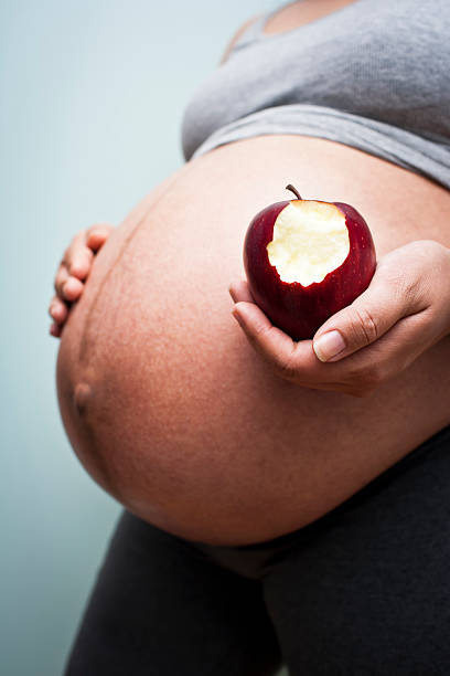 A pregnant woman practices healthy eating with an apple. Pregnant woman stands holding a partially eaten Macintosh apple apple with bite out of it stock pictures, royalty-free photos & images