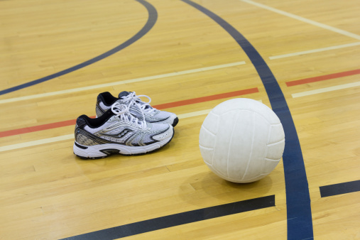 A volleyball with a pair of running shoes on a gym floor.  Focus is on the volleyball.