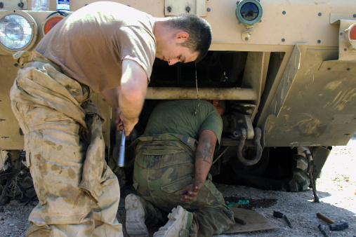 A U.S. Army mechanic hands tools to his buddy who is stripping parts off of a destroyed vehicle in Iraq