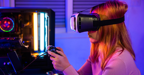 Excited woman virtual playing gamepad controller she celebrating victory at home, Gamer in VR headset glasses exploring metaverse play video game online with joystick on computer neon lights