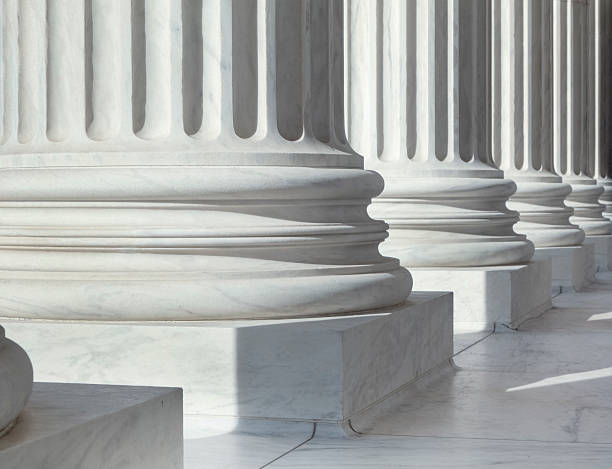 Column outside U.S. Supreme Court building An image of architectural detail from the United States Supreme Court building.  There are circular pillars visible in a row.  The pillars are white.  Each pillar has a block at the bottom, followed by three circular lines going all around them.  Vertical lines come next in an alternating pattern of sunken lines and protruding ones.  There are six pillars visible in the image, but only their lower parts can be seen.  The floor has white tiles.  There is sunlight coming from the front of the building, creating shadows behind the pillars. politics stock pictures, royalty-free photos & images