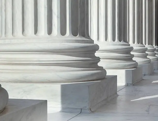 An image of architectural detail from the United States Supreme Court building.  There are circular pillars visible in a row.  The pillars are white.  Each pillar has a block at the bottom, followed by three circular lines going all around them.  Vertical lines come next in an alternating pattern of sunken lines and protruding ones.  There are six pillars visible in the image, but only their lower parts can be seen.  The floor has white tiles.  There is sunlight coming from the front of the building, creating shadows behind the pillars.