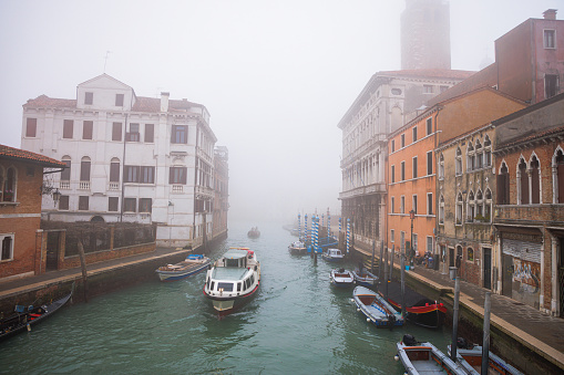 Secondary canal between residential buildings with small boats moored on the side, boat driving on water surface on a foggy winter day in Venice, Italy