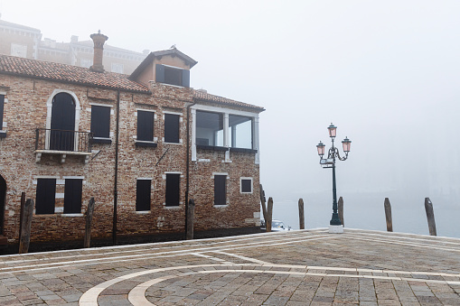 Stone paved square with old street light and house by the canal on a foggy winter day in Venice, Italy