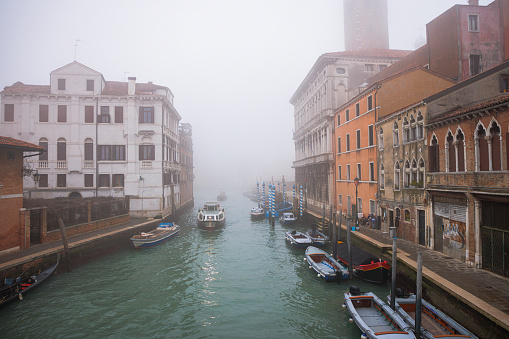 Secondary canal between residential buildings, small boats moored on the side, boat driving on water surface on a foggy day in Venice, Italy
