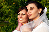 A happy charming bride in a white dress with a white veil and a bridesmaid with a pink veil hug gently and smile thoughtfully. Cheerful bachelorette party in the summer park. Close-up portrait.