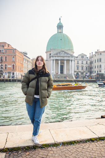 Beautiful teenage girl standing on pier by Grand Canale and looking at camera, church Santa Maria della Salute across the canal, enjoying her winter break in Venice, Italy