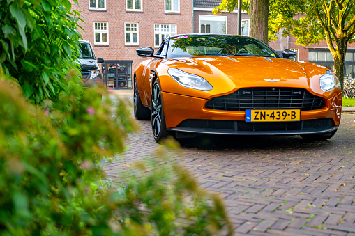 Aston Martin DB11 sports car in bright orange in the streets of Zwolle with people looking at the car. The Aston Martin DB11 is a luxury grand tourer unveiled in 2016 and available with V8 and V12 petrol engines.