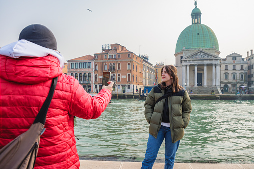 Beautiful teenage girl standing by Grand Canale while her father is taking a photo of her with smart phone, family enjoying vacation together in Venice, Italy