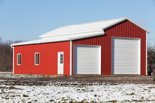 In early winter, construction is almost complete on this brand new, bright red and white corrugated metal warehouse/vehicle storage building on a rural road at the edge of a large apple orchard in western New York state, USA. Apple trees can be seen in the background.