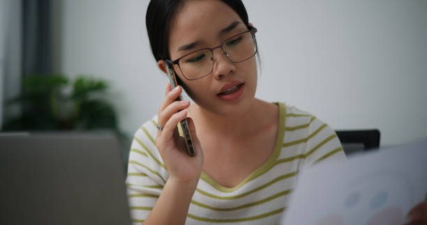 istock Young Asian woman wearing glasses freelance work holding a document while talking on mobile phone in home office room 1849238028
