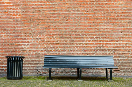 An empty bench and a garbage bin in front of a brick wall. Place your own object on the bench.See also