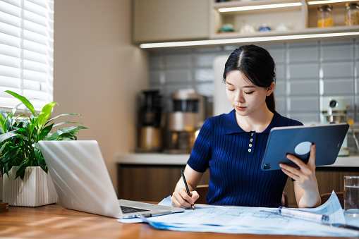 This is an Asian female designer, wearing casual clothing and sitting in the dining room at home. She has a blueprint in front of her, and she is working.