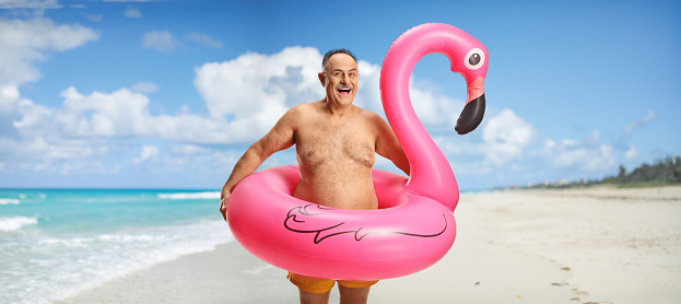 Happy mature man with a big inflatable flamingo rubber ring posing on a beach
