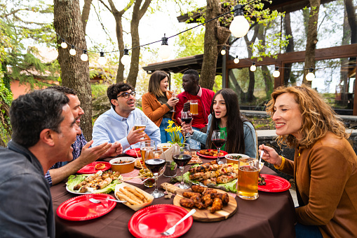 diverse friends having brunch on patio with bulb light in outdoor - multiracial young people toasting in home terrace with beer glasses and wine glasses in outside