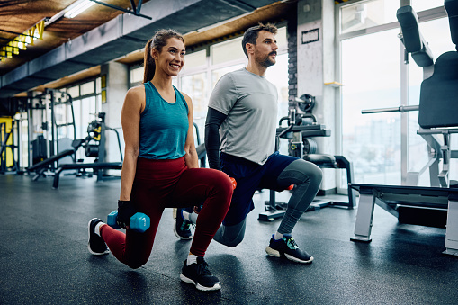 Athletic couple using dumbbells while exercising in lunge position in a gym. Copy space.